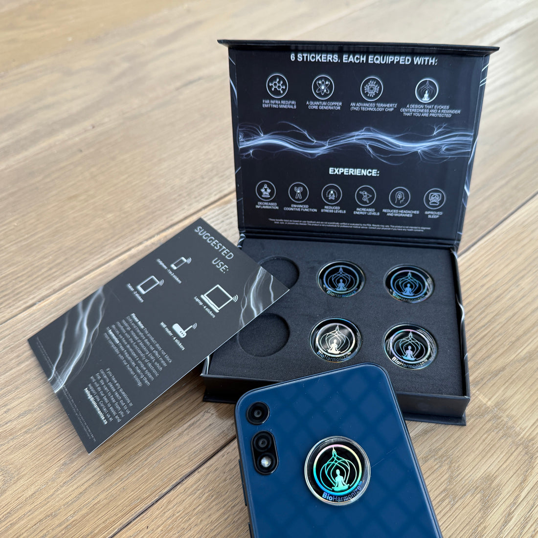 Open BioHarmonize EMF sticker box with six holographic stickers and a smartphone with one sticker applied to the back