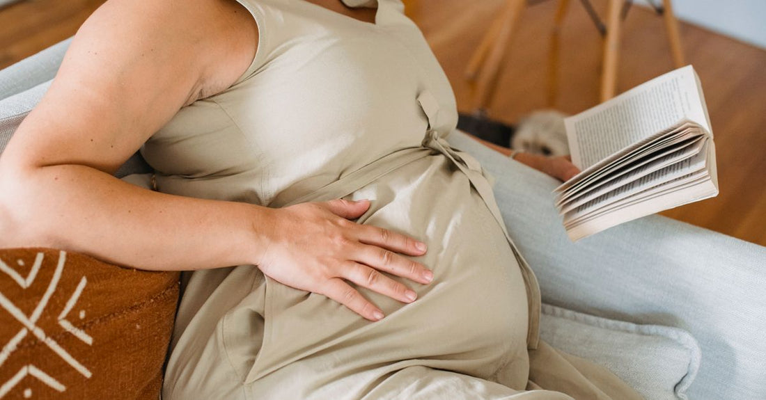 Pregnant woman in a beige dress reading a book on a sofa