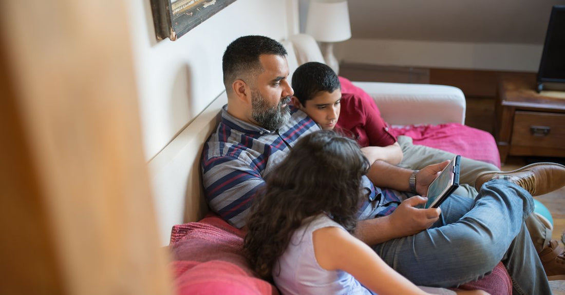 A father and his children relaxing on a couch using a digital tablet in a cozy home setting.